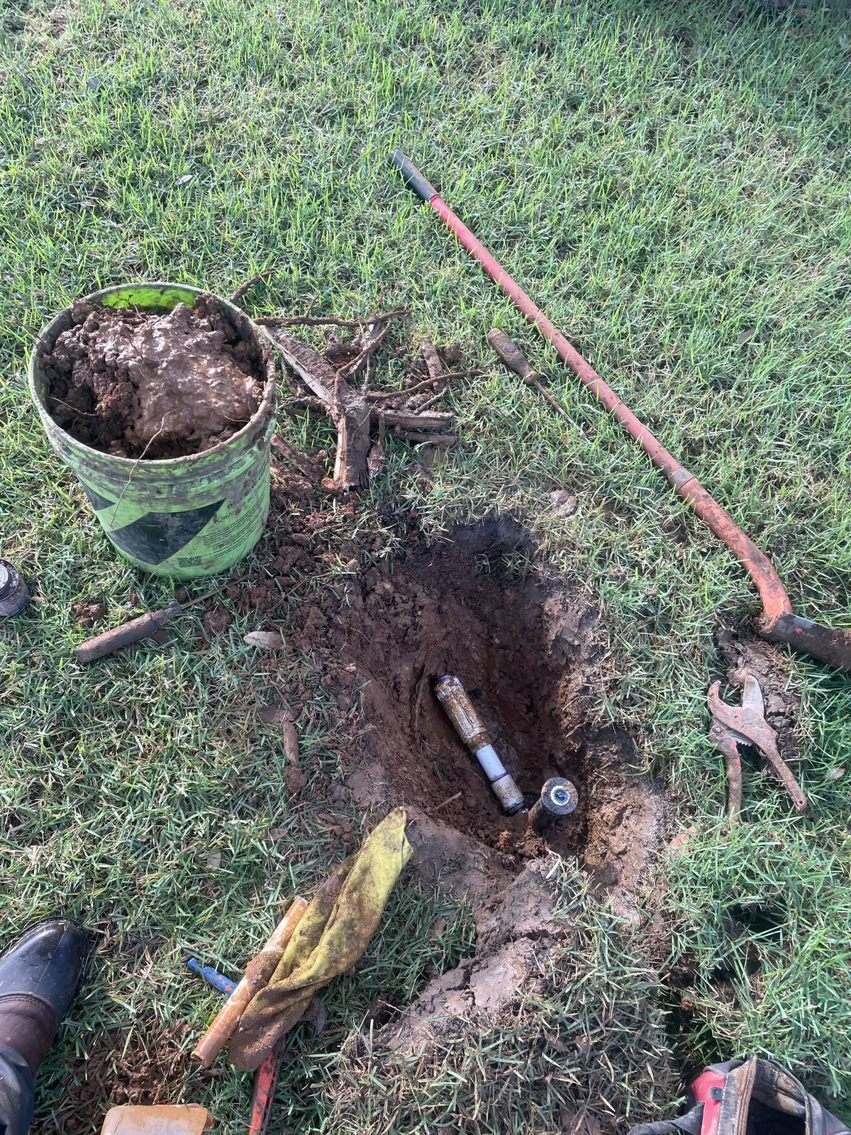 Hole dug to reach a sprinkler line in Grapevine, Texas, with a bucket of clay soil, cut tree roots and a pry bar beside it