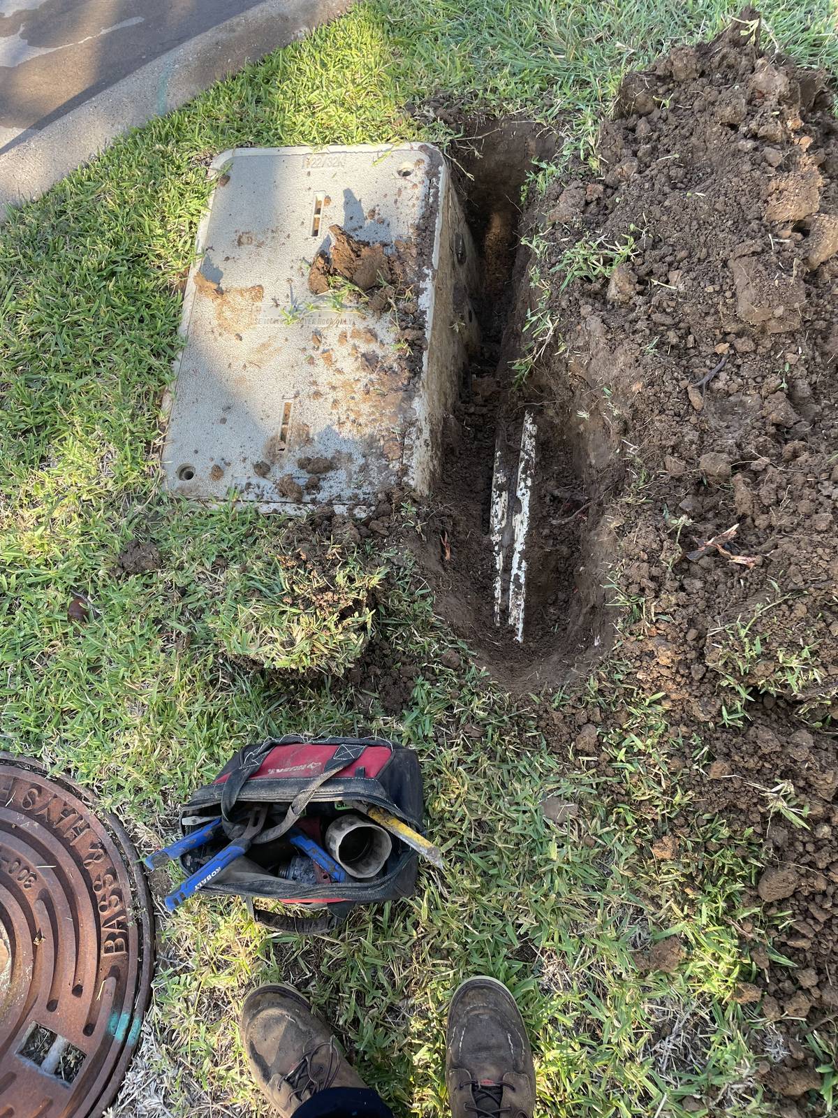 Trench dug beside a large concrete in-ground box near the curb in North Richland Hills, Texas to expose a leaking pipe, with a round meter lid in the corner